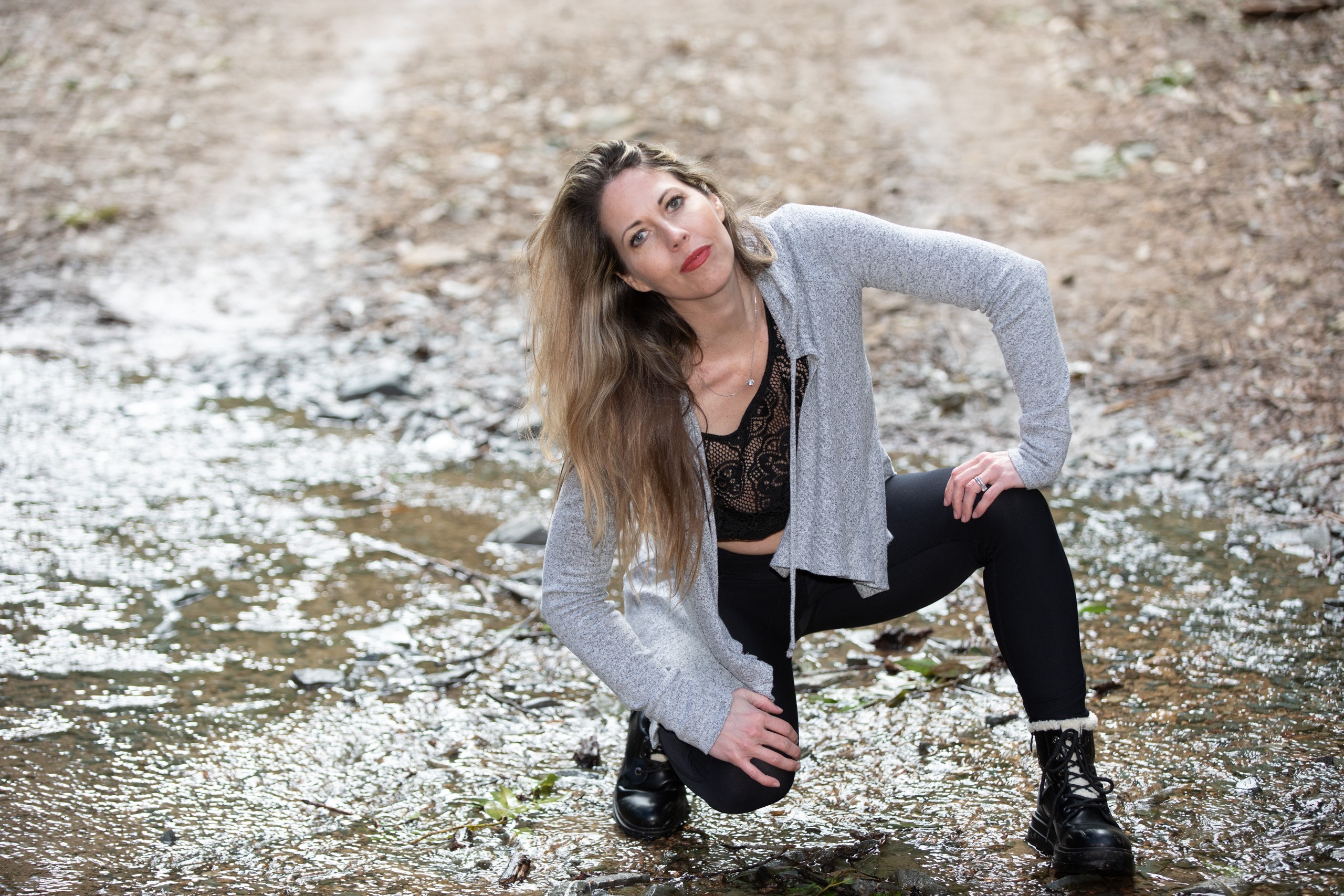 Woman in gray cardigan crouching by rocky stream, thoughtful expression
