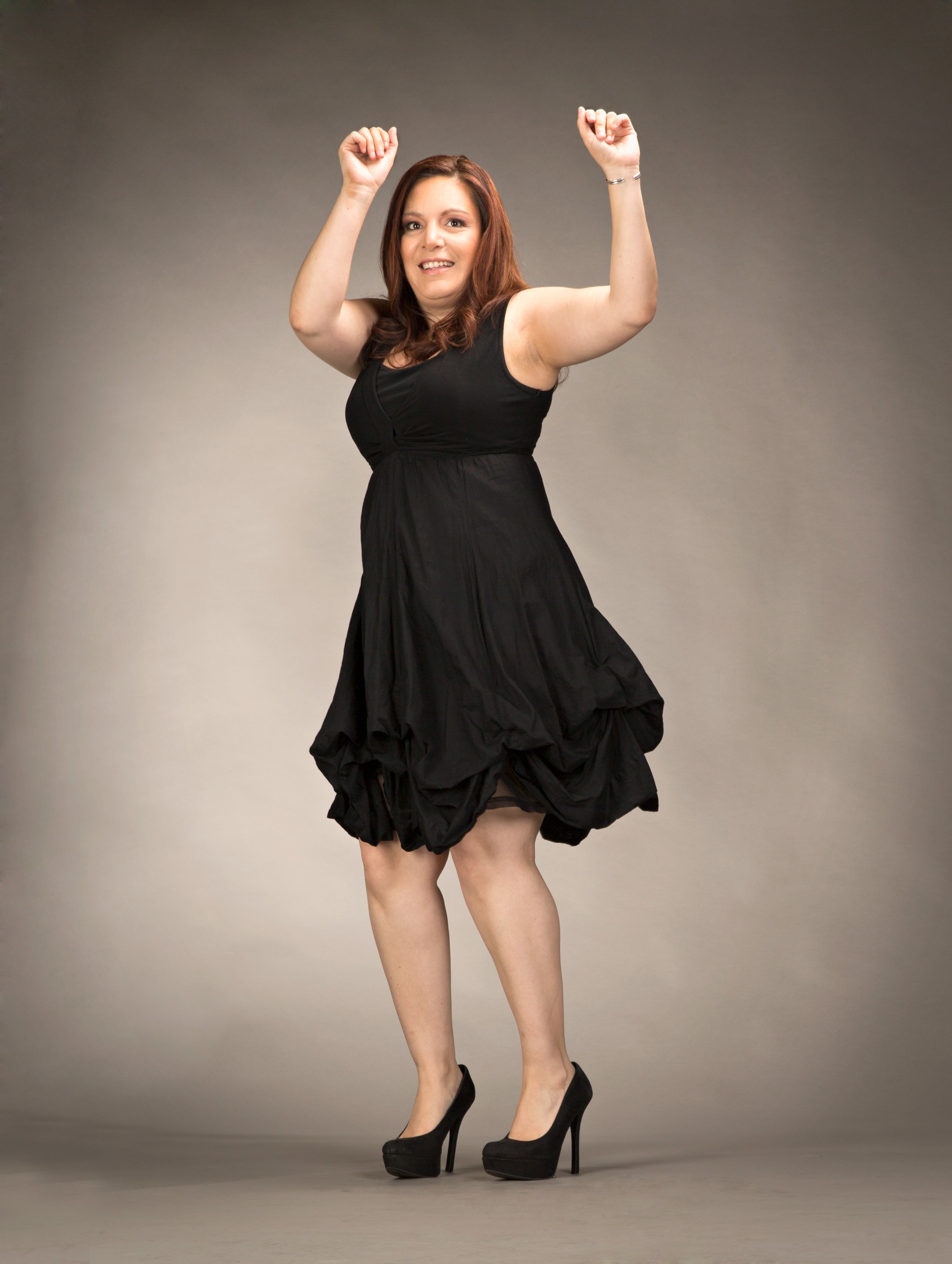 Woman in black dress celebrating with raised arms against neutral background